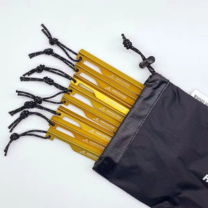 Les piquets de tente "Hikevictor - 15 cm" de couleur dorée, fabriqués en alliage d'aluminium léger, avec des cordons noirs, sont partiellement retirés d'une élégante housse de rangement noire.