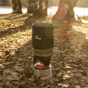 Un Fire Maple Fixed Star 1 réchaud de camping / brûleur à gaz système de cuisson ultra efficace 550g avec un pot noir est installé en plein air sur des feuilles mortes, entouré d'arbres et de la lumière du soleil en arrière-plan.