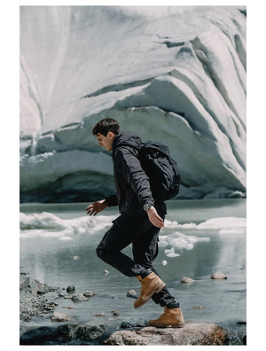 Une personne portant la veste de randonnée coupe-vent avec traitement hydrofuge pour homme Simwood « SIMWOOD - SM130288 », un pantalon noir et des bottes marron marche sur des rochers près d'un lac glaciaire avec des formations de glace en arrière-plan.