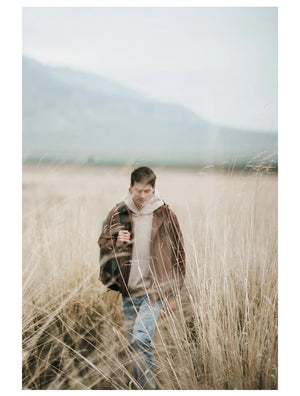 Une personne portant une Veste de randonnée coupe-vent avec traitement hydrofuge pour homme Simwood « SIMWOOD - SM130288 » et portant un sac marche dans les hautes herbes d'un champ avec des montagnes en arrière-plan.