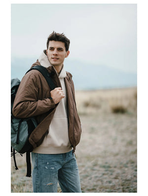 Une personne se tient debout à l'extérieur, vêtue d'une veste Simwood marron coupe-vent et imperméable (SIMWOOD - SM130288), d'un sweat à capuche beige et d'un jean, avec un sac à dos noir sur une épaule. L'arrière-plan montre un champ ouvert sous un ciel couvert.