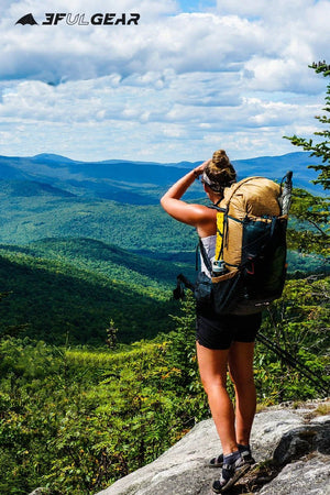 Une personne avec un sac à dos 3F UL GEAR Qi Dian Pro se tient sur un rebord rocheux, surplombant une vallée boisée et des montagnes lointaines sous un ciel partiellement nuageux.
