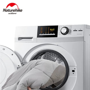 Une machine à laver à chargement frontal avec une porte ouverte et une couette partiellement à l'intérieur met en valeur les réglages du panneau de commande. Le logo Naturehike en haut à gauche suggère des matériaux en nylon durables, idéaux pour le "Naturehike - NH21MSD03" Sac de couchage en coton imperméable pour l'hiver.
