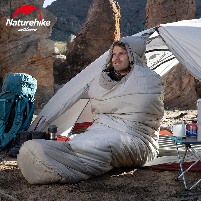 Une personne est confortablement assise dans une tente avec le logo Naturehike, nichée à l'intérieur d'un sac de couchage en coton imperméable pour l'hiver « Naturehike - NH21MSD03 », entourée d'un terrain rocheux. Le tissu en nylon durable offre une protection contre les éléments.
