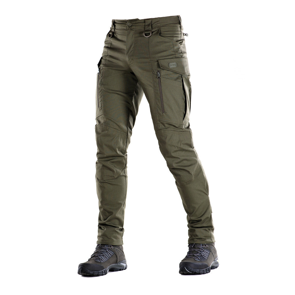 Un homme portant le pantalon M-TAC "M-Tac Conquistador Gen. I Flex" cargo tactique militaire résistant à l'usure et à l'eau, avec des poches utilitaires, et des chaussures noires à lacets, se tient les jambes écartées sur un fond blanc.