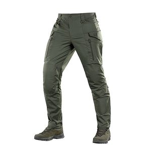 Un homme portant le pantalon M-TAC "M-Tac Conquistador Gen. I Flex" cargo tactique militaire résistant à l'usure et à l'eau, avec des poches utilitaires, et des chaussures noires à lacets, se tient les jambes écartées sur un fond blanc.