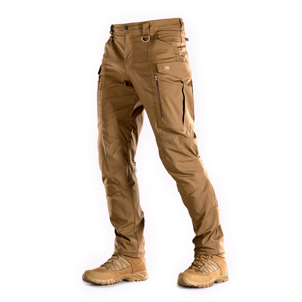 Un homme portant le pantalon M-TAC "M-Tac Conquistador Gen. I Flex" cargo tactique militaire résistant à l'usure et à l'eau, avec des poches utilitaires, et des chaussures noires à lacets, se tient les jambes écartées sur un fond blanc.