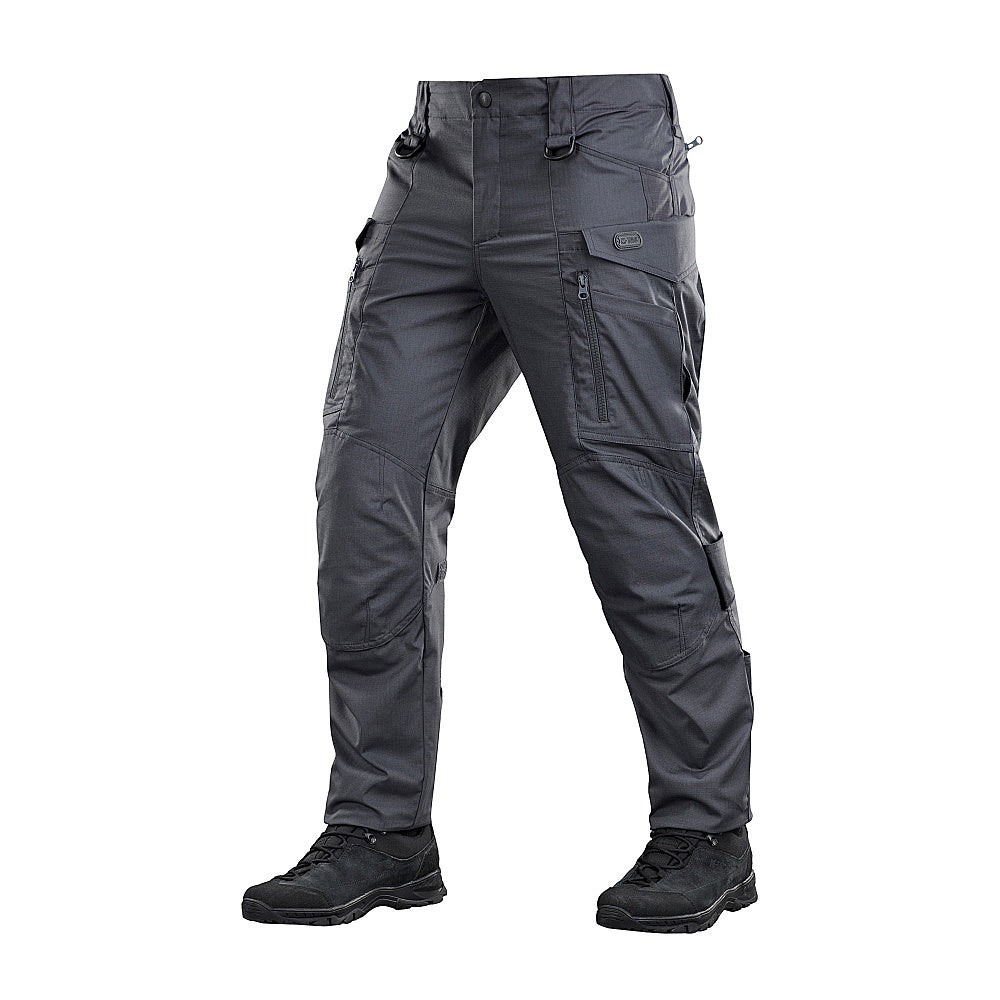 Un homme portant le pantalon M-TAC "M-Tac Conquistador Gen. I Flex" cargo tactique militaire résistant à l'usure et à l'eau, avec des poches utilitaires, et des chaussures noires à lacets, se tient les jambes écartées sur un fond blanc.