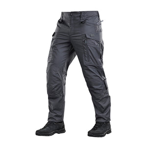 Un homme portant le pantalon M-TAC "M-Tac Conquistador Gen. I Flex" cargo tactique militaire résistant à l'usure et à l'eau, avec des poches utilitaires, et des chaussures noires à lacets, se tient les jambes écartées sur un fond blanc.