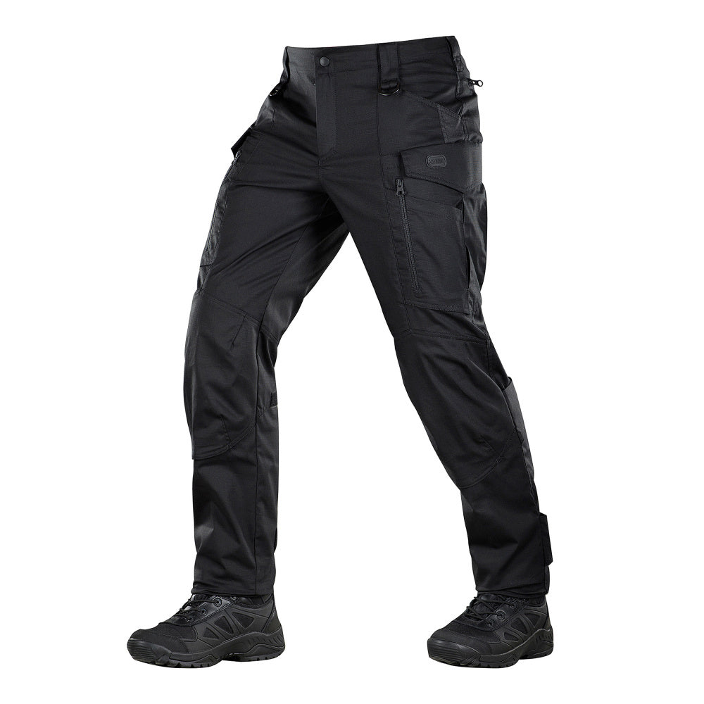 Un homme portant le pantalon M-TAC "M-Tac Conquistador Gen. I Flex" cargo tactique militaire résistant à l'usure et à l'eau, avec des poches utilitaires, et des chaussures noires à lacets, se tient les jambes écartées sur un fond blanc.