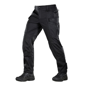 Un homme portant le pantalon M-TAC "M-Tac Conquistador Gen. I Flex" cargo tactique militaire résistant à l'usure et à l'eau, avec des poches utilitaires, et des chaussures noires à lacets, se tient les jambes écartées sur un fond blanc.