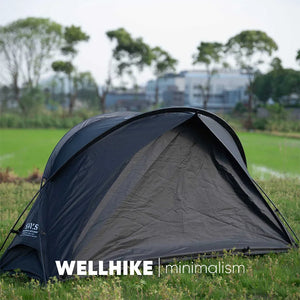 A Lit de camp 2 en 1 avec tente simple-paroi "Wellhike Minimalism - Yunchi SWS" de Wellhike Minimalism est installé sur l'herbe, avec des tables pliantes à proximité. Le texte "WELLHIKE minimalism" apparaît en bas de l'image.