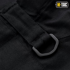 Un homme portant le pantalon M-TAC "M-Tac Conquistador Gen. I Flex" cargo tactique militaire résistant à l'usure et à l'eau, avec des poches utilitaires, et des chaussures noires à lacets, se tient les jambes écartées sur un fond blanc.