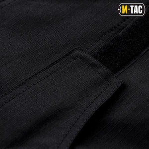 Un homme portant le pantalon M-TAC "M-Tac Conquistador Gen. I Flex" cargo tactique militaire résistant à l'usure et à l'eau, avec des poches utilitaires, et des chaussures noires à lacets, se tient les jambes écartées sur un fond blanc.