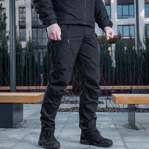 Un homme portant le pantalon M-TAC "M-Tac Conquistador Gen. I Flex" cargo tactique militaire résistant à l'usure et à l'eau, avec des poches utilitaires, et des chaussures noires à lacets, se tient les jambes écartées sur un fond blanc.