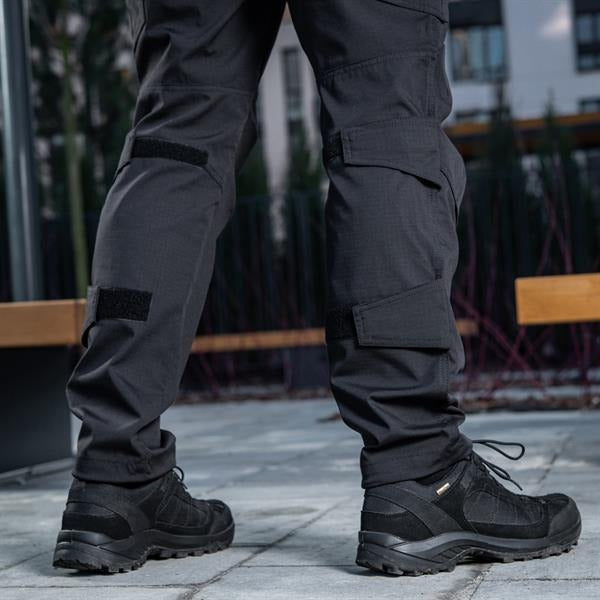 Un homme portant le pantalon M-TAC "M-Tac Conquistador Gen. I Flex" cargo tactique militaire résistant à l'usure et à l'eau, avec des poches utilitaires, et des chaussures noires à lacets, se tient les jambes écartées sur un fond blanc.