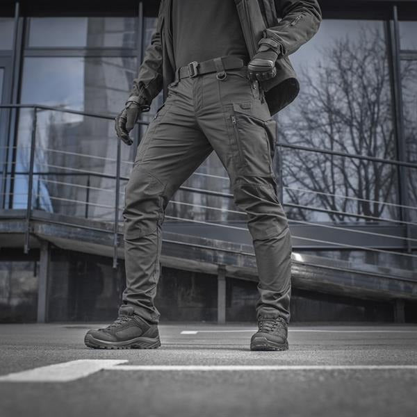 Un homme portant le pantalon M-TAC "M-Tac Conquistador Gen. I Flex" cargo tactique militaire résistant à l'usure et à l'eau, avec des poches utilitaires, et des chaussures noires à lacets, se tient les jambes écartées sur un fond blanc.