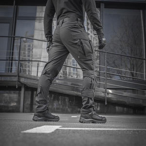 Un homme portant le pantalon M-TAC "M-Tac Conquistador Gen. I Flex" cargo tactique militaire résistant à l'usure et à l'eau, avec des poches utilitaires, et des chaussures noires à lacets, se tient les jambes écartées sur un fond blanc.