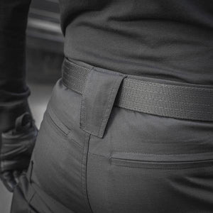 Un homme portant le pantalon M-TAC "M-Tac Conquistador Gen. I Flex" cargo tactique militaire résistant à l'usure et à l'eau, avec des poches utilitaires, et des chaussures noires à lacets, se tient les jambes écartées sur un fond blanc.