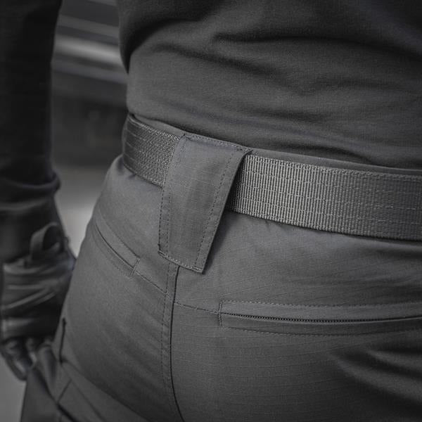 Un homme portant le pantalon M-TAC "M-Tac Conquistador Gen. I Flex" cargo tactique militaire résistant à l'usure et à l'eau, avec des poches utilitaires, et des chaussures noires à lacets, se tient les jambes écartées sur un fond blanc.