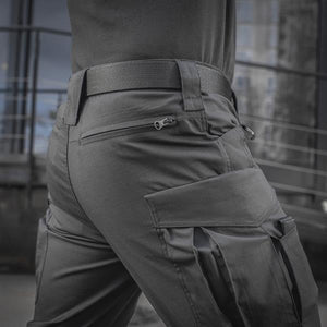 Un homme portant le pantalon M-TAC "M-Tac Conquistador Gen. I Flex" cargo tactique militaire résistant à l'usure et à l'eau, avec des poches utilitaires, et des chaussures noires à lacets, se tient les jambes écartées sur un fond blanc.