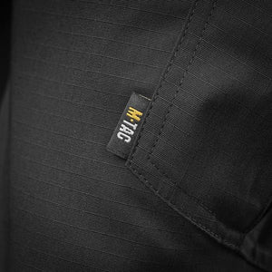 Un homme portant le pantalon M-TAC "M-Tac Conquistador Gen. I Flex" cargo tactique militaire résistant à l'usure et à l'eau, avec des poches utilitaires, et des chaussures noires à lacets, se tient les jambes écartées sur un fond blanc.