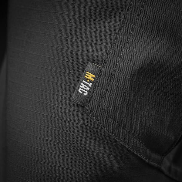 Un homme portant le pantalon M-TAC "M-Tac Conquistador Gen. I Flex" cargo tactique militaire résistant à l'usure et à l'eau, avec des poches utilitaires, et des chaussures noires à lacets, se tient les jambes écartées sur un fond blanc.