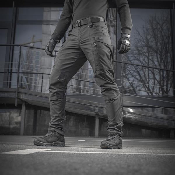 Un homme portant le pantalon M-TAC "M-Tac Conquistador Gen. I Flex" cargo tactique militaire résistant à l'usure et à l'eau, avec des poches utilitaires, et des chaussures noires à lacets, se tient les jambes écartées sur un fond blanc.