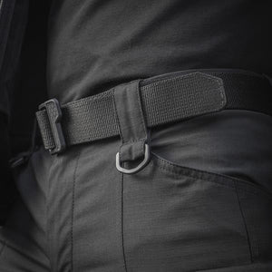 Un homme portant le pantalon M-TAC "M-Tac Conquistador Gen. I Flex" cargo tactique militaire résistant à l'usure et à l'eau, avec des poches utilitaires, et des chaussures noires à lacets, se tient les jambes écartées sur un fond blanc.