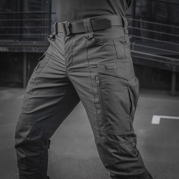 Un homme portant le pantalon M-TAC "M-Tac Conquistador Gen. I Flex" cargo tactique militaire résistant à l'usure et à l'eau, avec des poches utilitaires, et des chaussures noires à lacets, se tient les jambes écartées sur un fond blanc.