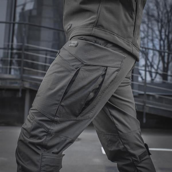 Un homme portant le pantalon M-TAC "M-Tac Conquistador Gen. I Flex" cargo tactique militaire résistant à l'usure et à l'eau, avec des poches utilitaires, et des chaussures noires à lacets, se tient les jambes écartées sur un fond blanc.