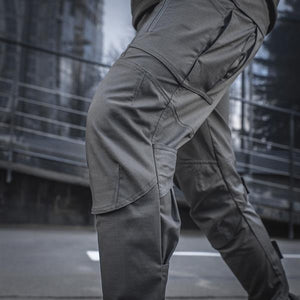 Un homme portant le pantalon M-TAC "M-Tac Conquistador Gen. I Flex" cargo tactique militaire résistant à l'usure et à l'eau, avec des poches utilitaires, et des chaussures noires à lacets, se tient les jambes écartées sur un fond blanc.