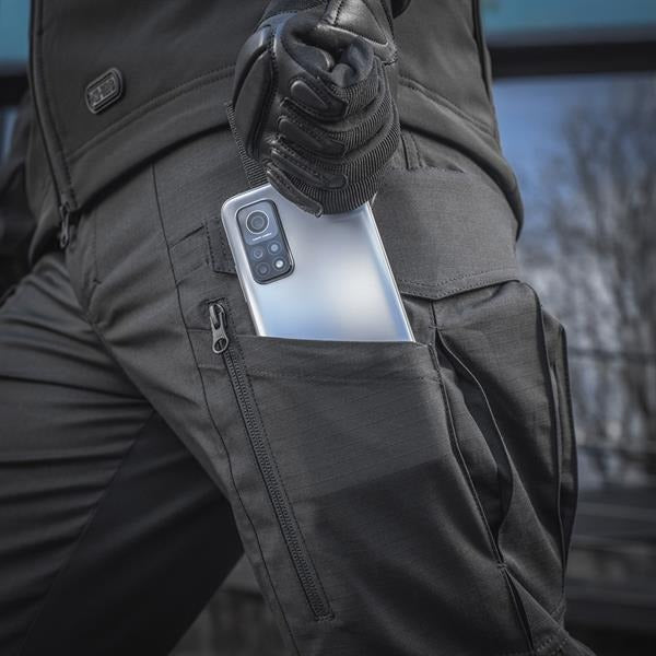 Un homme portant le pantalon M-TAC "M-Tac Conquistador Gen. I Flex" cargo tactique militaire résistant à l'usure et à l'eau, avec des poches utilitaires, et des chaussures noires à lacets, se tient les jambes écartées sur un fond blanc.