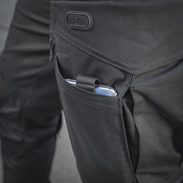 Un homme portant le pantalon M-TAC "M-Tac Conquistador Gen. I Flex" cargo tactique militaire résistant à l'usure et à l'eau, avec des poches utilitaires, et des chaussures noires à lacets, se tient les jambes écartées sur un fond blanc.