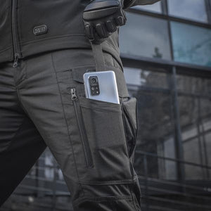 Un homme portant le pantalon M-TAC "M-Tac Conquistador Gen. I Flex" cargo tactique militaire résistant à l'usure et à l'eau, avec des poches utilitaires, et des chaussures noires à lacets, se tient les jambes écartées sur un fond blanc.