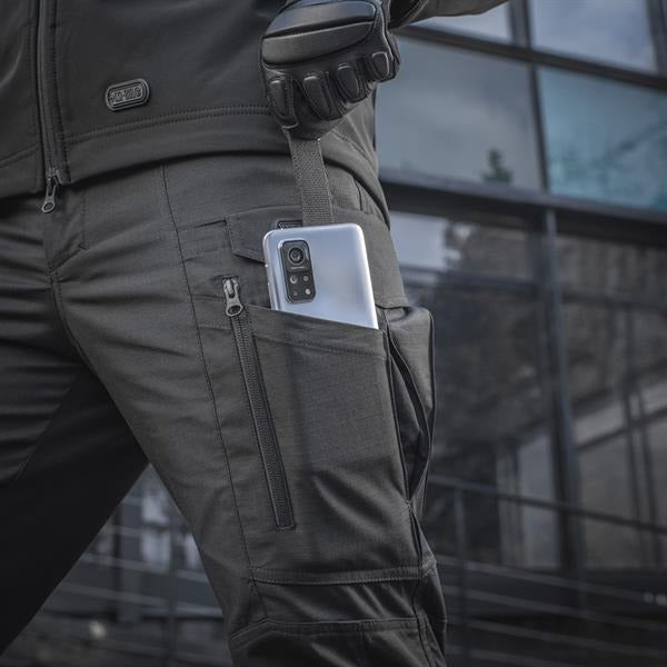 Un homme portant le pantalon M-TAC "M-Tac Conquistador Gen. I Flex" cargo tactique militaire résistant à l'usure et à l'eau, avec des poches utilitaires, et des chaussures noires à lacets, se tient les jambes écartées sur un fond blanc.