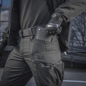 Un homme portant le pantalon M-TAC "M-Tac Conquistador Gen. I Flex" cargo tactique militaire résistant à l'usure et à l'eau, avec des poches utilitaires, et des chaussures noires à lacets, se tient les jambes écartées sur un fond blanc.