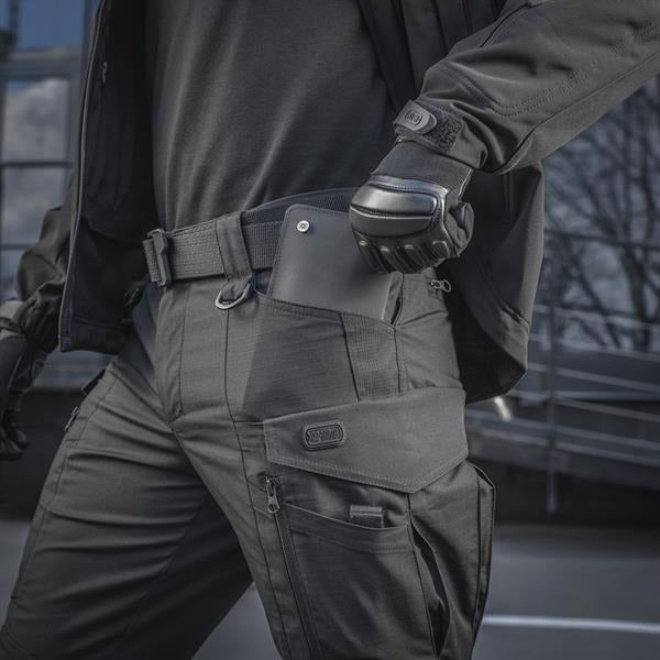 Un homme portant le pantalon M-TAC "M-Tac Conquistador Gen. I Flex" cargo tactique militaire résistant à l'usure et à l'eau, avec des poches utilitaires, et des chaussures noires à lacets, se tient les jambes écartées sur un fond blanc.