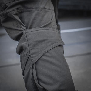 Un homme portant le pantalon M-TAC "M-Tac Conquistador Gen. I Flex" cargo tactique militaire résistant à l'usure et à l'eau, avec des poches utilitaires, et des chaussures noires à lacets, se tient les jambes écartées sur un fond blanc.