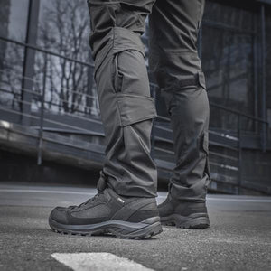 Un homme portant le pantalon M-TAC "M-Tac Conquistador Gen. I Flex" cargo tactique militaire résistant à l'usure et à l'eau, avec des poches utilitaires, et des chaussures noires à lacets, se tient les jambes écartées sur un fond blanc.