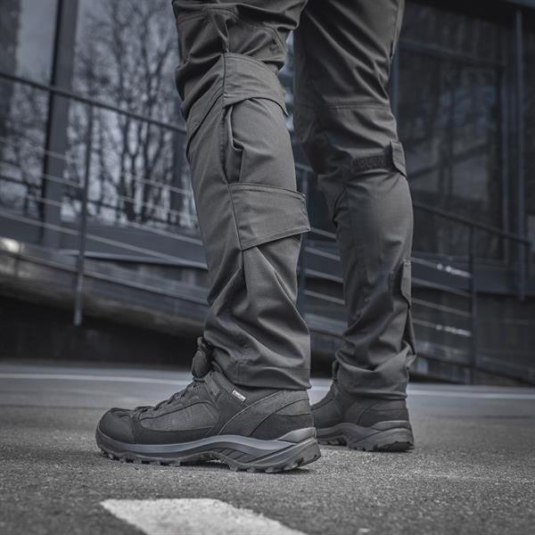 Un homme portant le pantalon M-TAC "M-Tac Conquistador Gen. I Flex" cargo tactique militaire résistant à l'usure et à l'eau, avec des poches utilitaires, et des chaussures noires à lacets, se tient les jambes écartées sur un fond blanc.