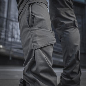 Un homme portant le pantalon M-TAC "M-Tac Conquistador Gen. I Flex" cargo tactique militaire résistant à l'usure et à l'eau, avec des poches utilitaires, et des chaussures noires à lacets, se tient les jambes écartées sur un fond blanc.
