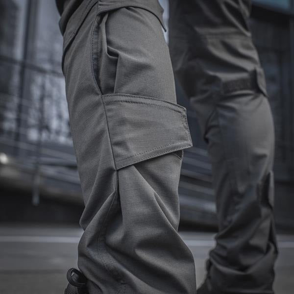 Un homme portant le pantalon M-TAC "M-Tac Conquistador Gen. I Flex" cargo tactique militaire résistant à l'usure et à l'eau, avec des poches utilitaires, et des chaussures noires à lacets, se tient les jambes écartées sur un fond blanc.